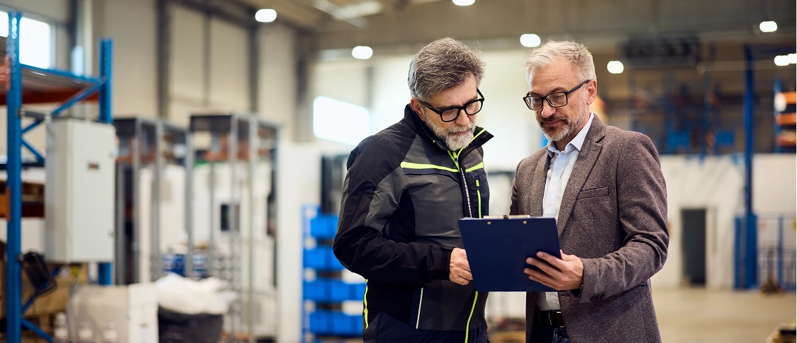 Two men standing in a warehouse discuss information on a clipboard; one wears a business suit, the other wears a work jacket. Shelving and equipment are visible in the background.